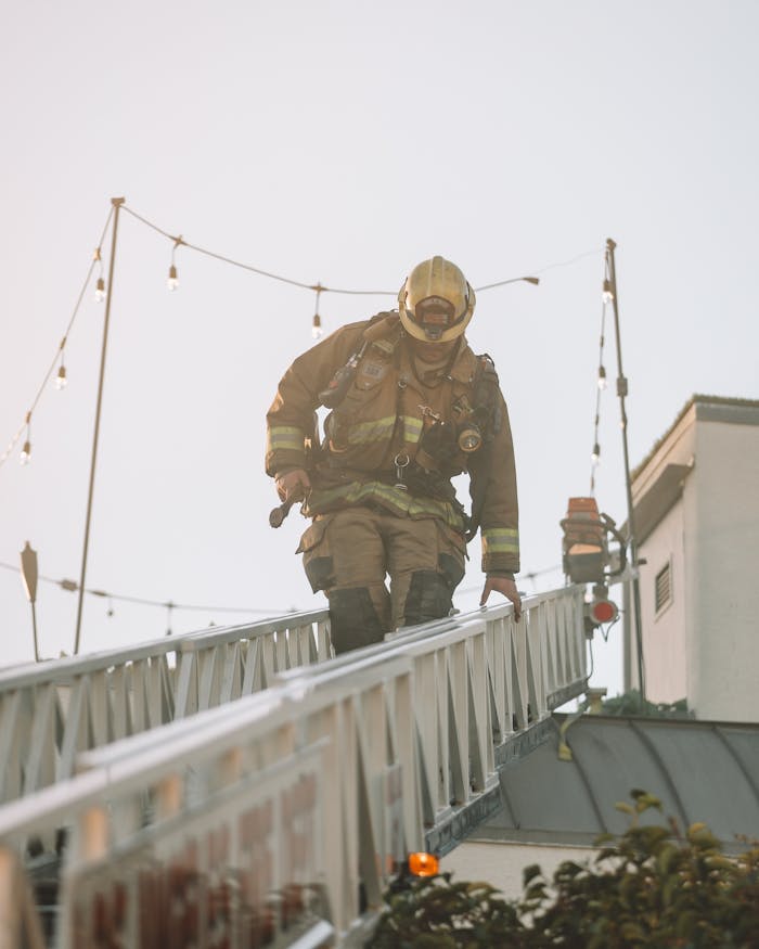 Firefighter in full gear climbs down a ladder outdoors surrounded by string lights.
